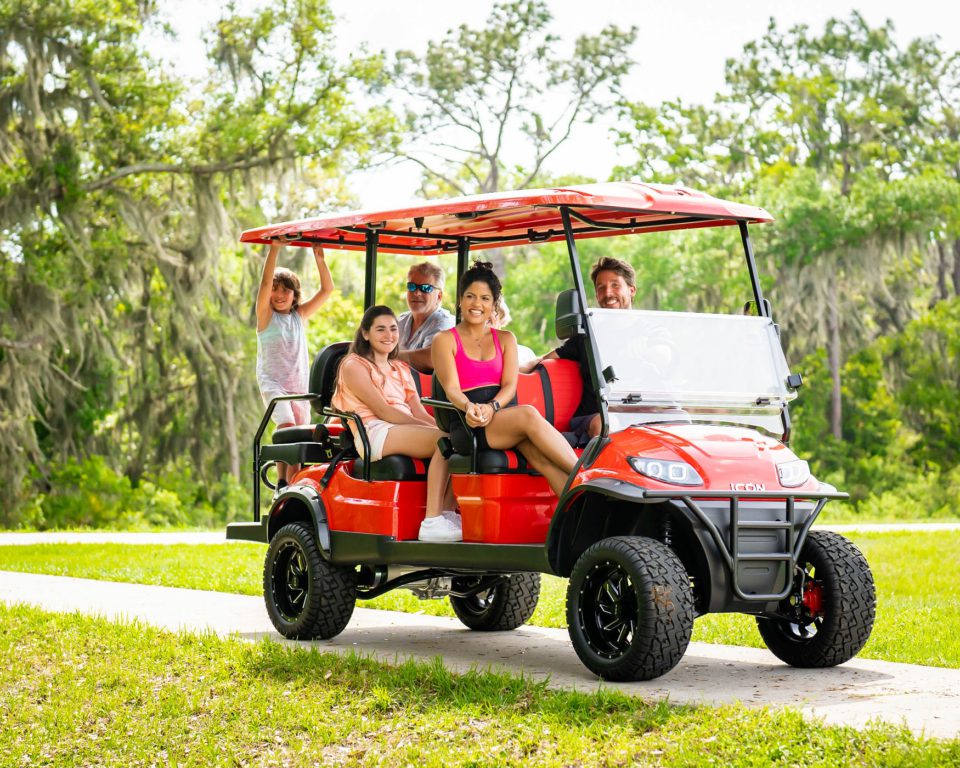 A family of 5 on a golf cart on a paved roadway with trees in the background.