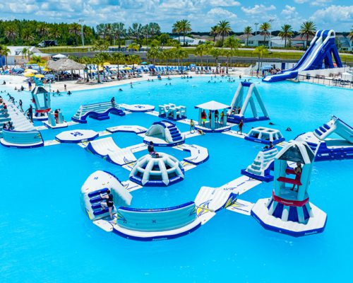 Inflatable obstacle course on top of clear blue water with climbing wall, bridges, slides, igloos, and various obstacles with kids in life vests playing. Beach in the background with guests lounging on beach chairs.