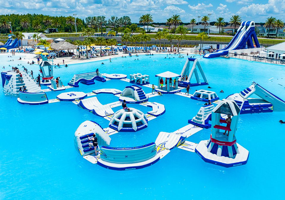 Inflatable obstacle course on top of clear blue water with climbing wall, bridges, slides, igloos, and various obstacles with kids in life vests playing. Beach in the background with guests lounging on beach chairs.